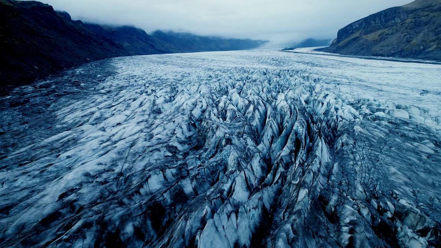 An expansive aerial view of a textured glacier with deep blue and white ridges, nestled between dark, misty mountains under an overcast sky.